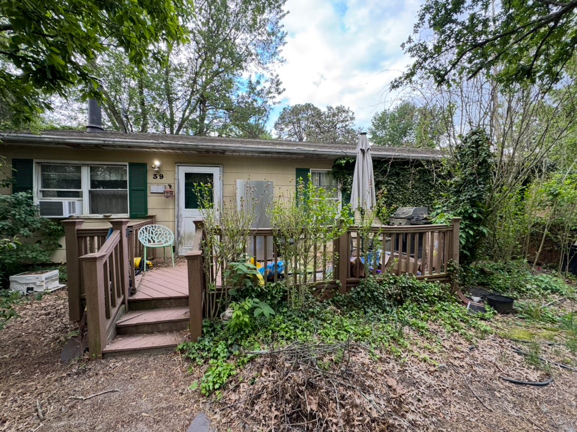 Overgrown single-story Howell, New Jersey home with front deck, mature trees, and exterior in need of cleanup and repairs.